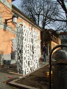 Große weiße Metallskulptur mit deutschen Wörtern vor einem gelben Gebäude, Mann arbeitet im Hintergrund